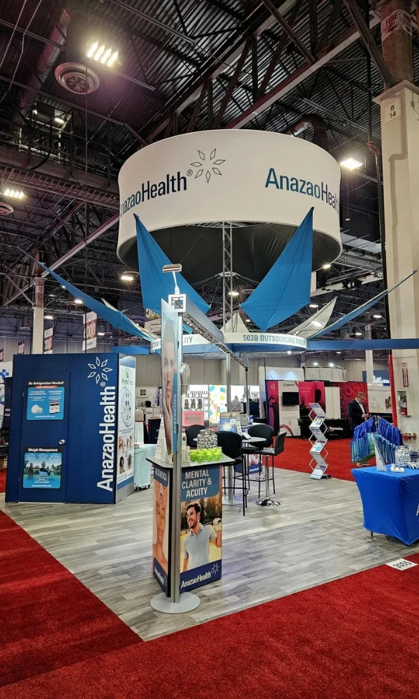 AnazaoHealth trade show booth with circular hanging sign, branded towers, counters, and seating on wood-look flooring by Suncoast Displays